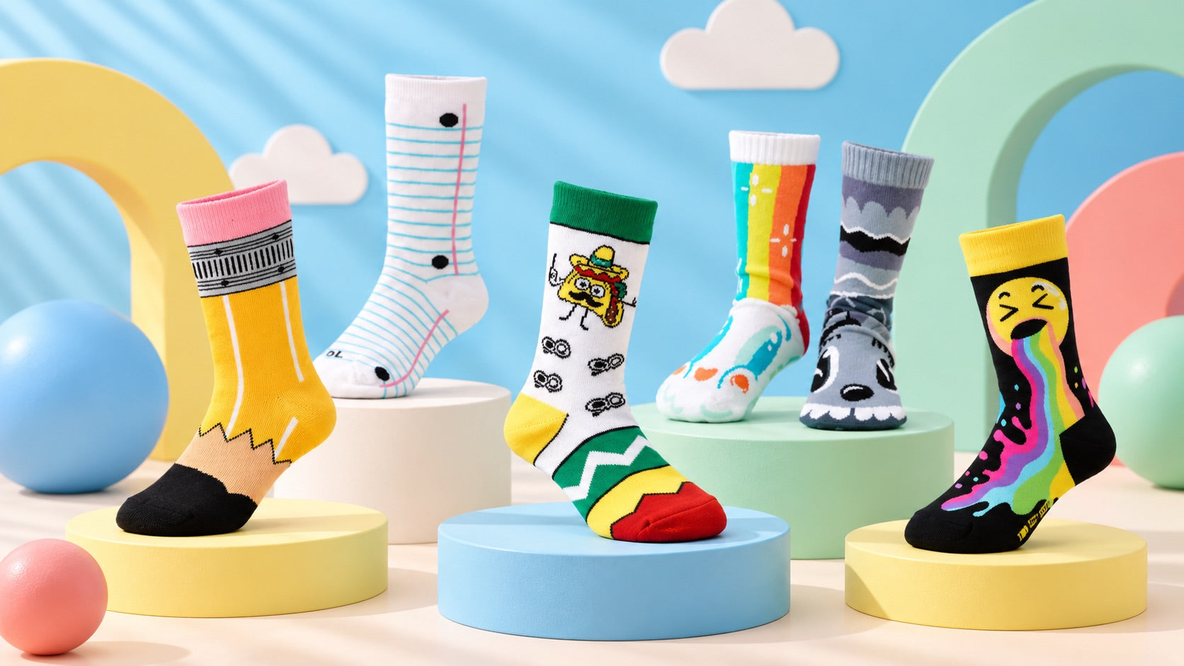 Six kids' Motsu socks on pastel pedestals against a blue sky backdrop with clouds: pencil, notebook paper, taco, rainbow, storm cloud and rainbow-tongue emoji