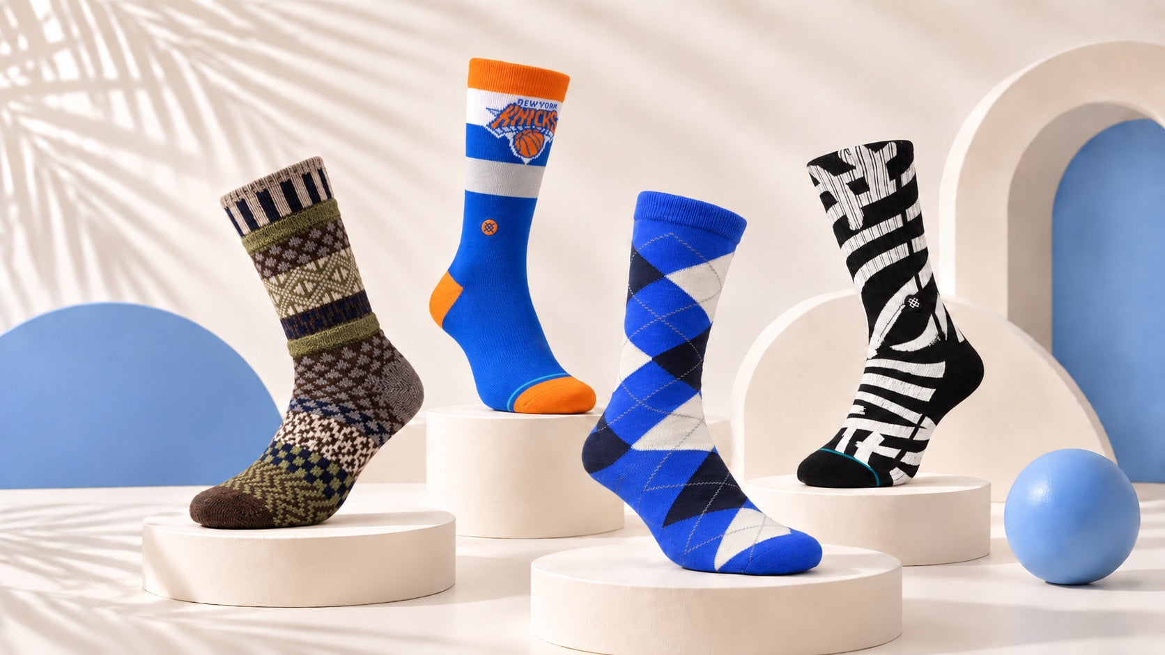 Four men's Motsu socks on cream pedestals against a cream and blue studio backdrop: patterned wool, New York Knicks, blue argyle and black and white graphic