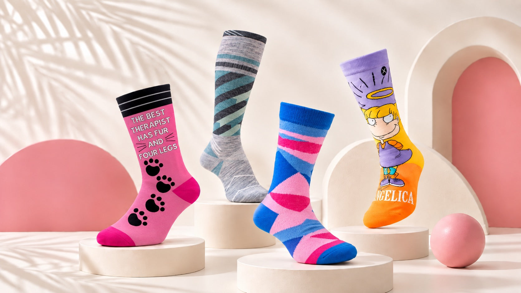 Four women's Motsu socks on cream pedestals against a cream and pink studio backdrop: a pink paw-print slogan sock, a grey striped knee-high, a pink and blue geometric sock, and an Angelica cartoon sock